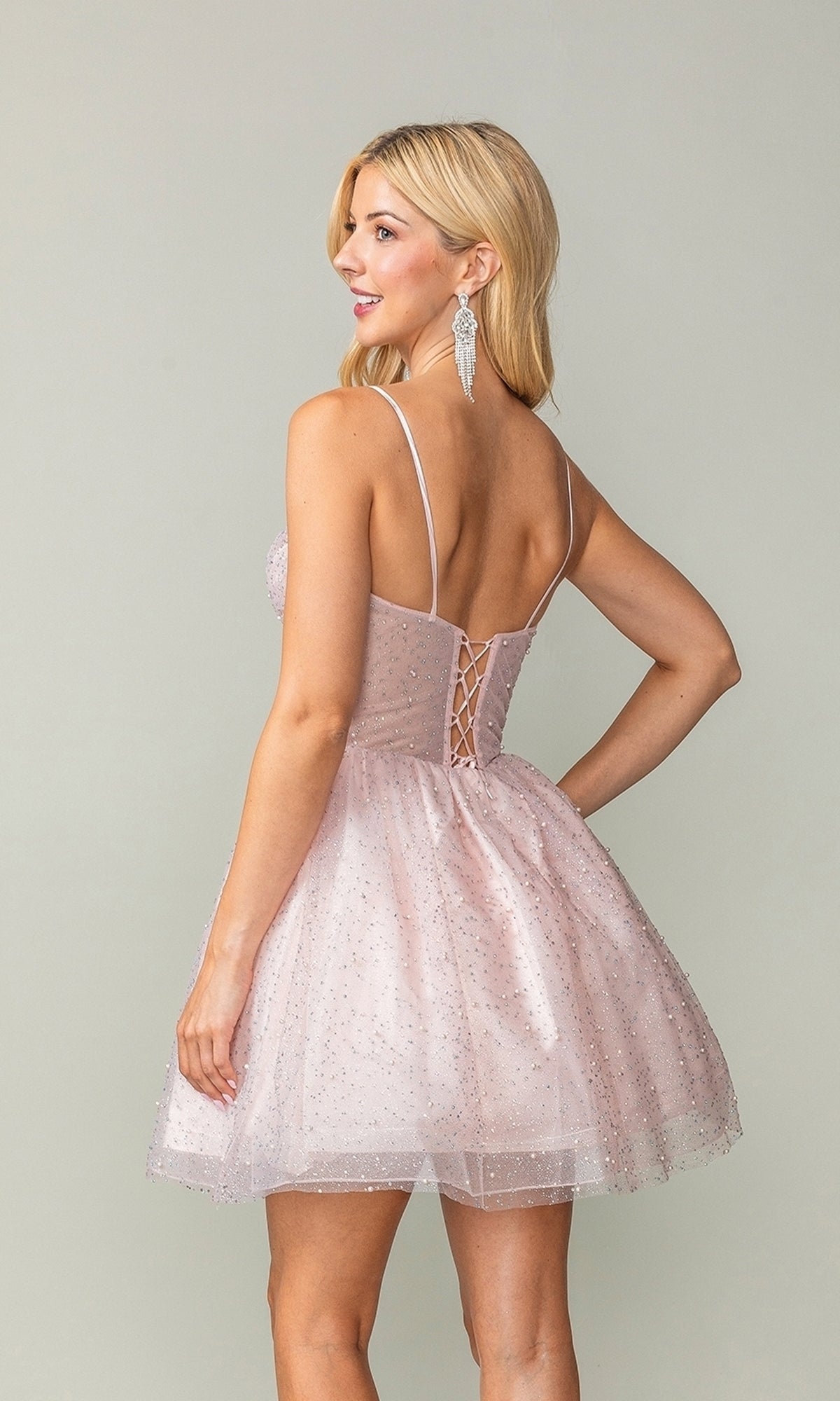 A woman in the Short Glitter Fit-and-Flare Homecoming Dress 3389, featuring light pink sparkly fabric, thin straps, a lace-up back, and a sleeveless fit-and-flare design, stands facing away and looking over her shoulder.